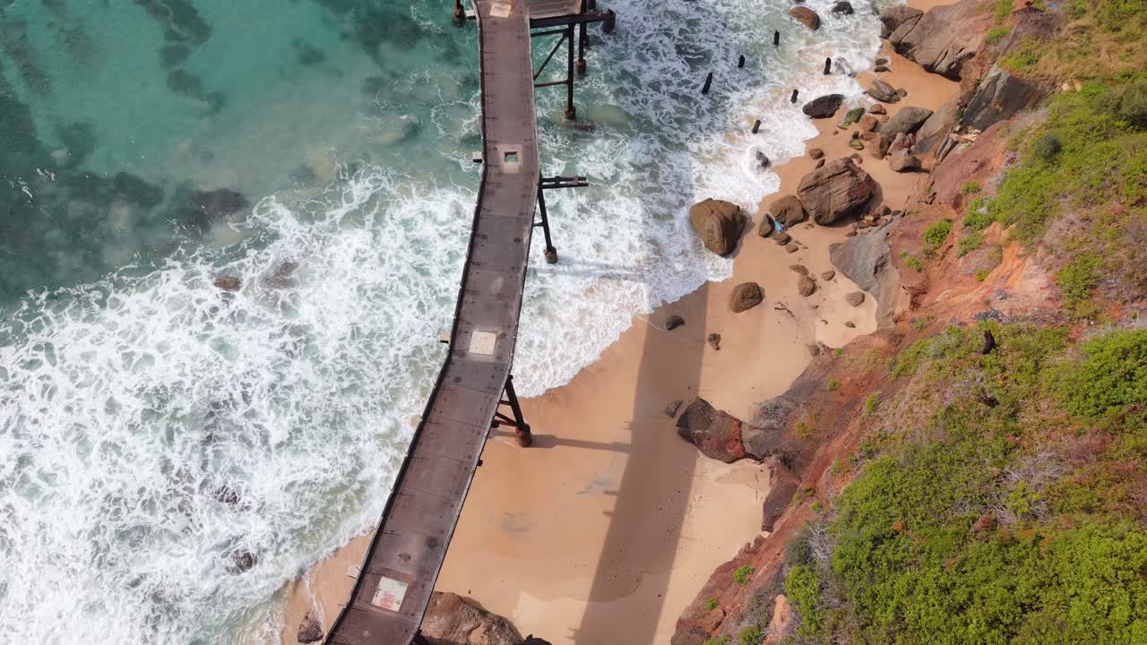 Overhead aerial of waves crashing on Catherine Hill Bay shoreline in New South Wales below metal structure