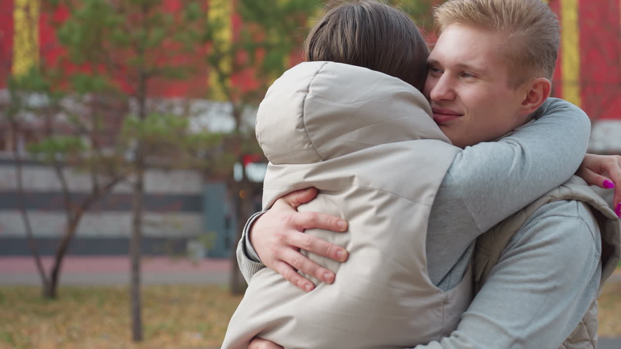 Side view of friends dressed in same outfit approaching each other with warm smiles and embracing in cheerful hug as autumn trees sway gently with wind and building background
