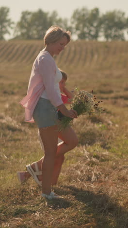 mother and daughter walking together in vast farmland under sunny sky with mother holding fresh flower bouquet in her right hand, surrounded by open field and distant trees