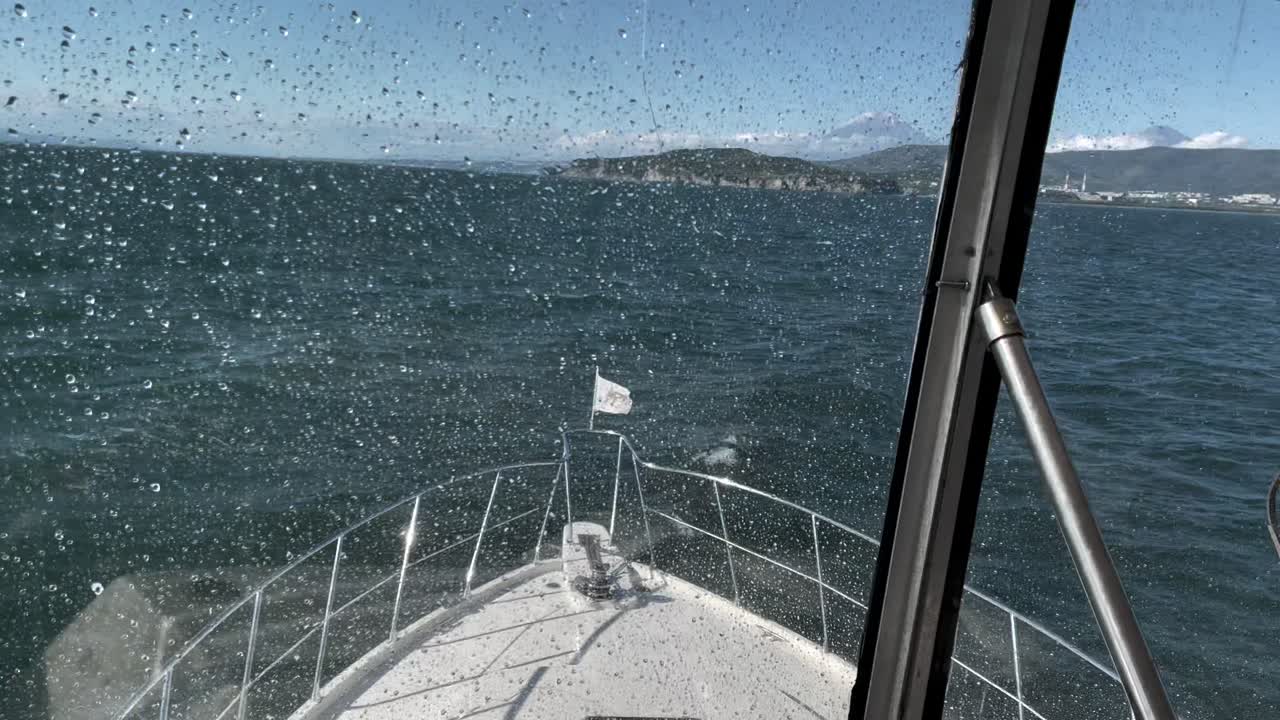 POV von einem fahrenden Motorboot mit Gischt auf der Windschutzscheibe, das über unruhiges Wasser auf eine zerklüftete Küste und ferne Berge in Kamtschatka, Russland, zusteuert