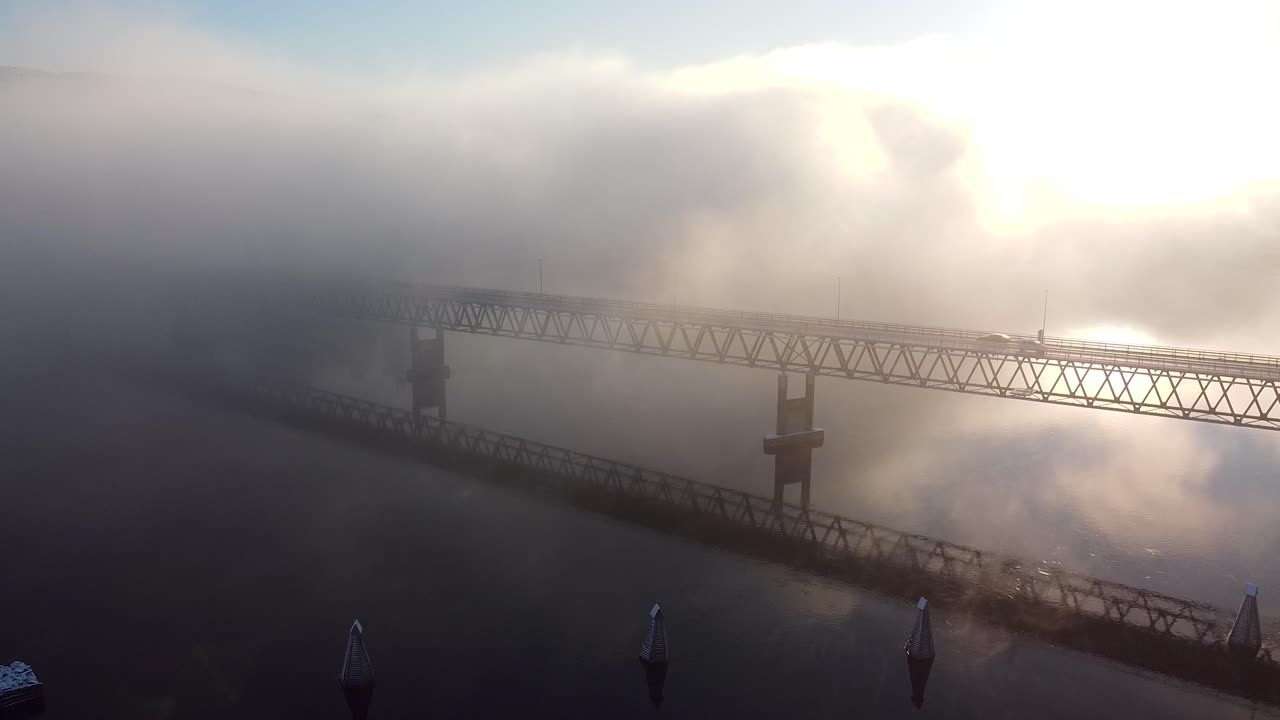 A car drives across a mist-covered bridge in Norway as morning light cuts through the dense fog