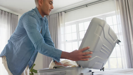 Happy-biracial-man-packing-suitcase-in-bright-bedroom