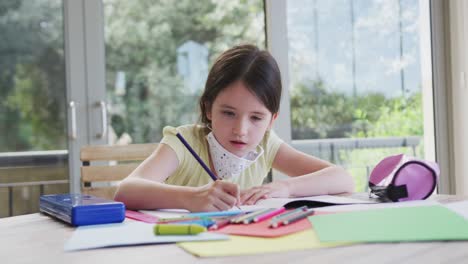 Caucasian-girl-spending-time-at-home-with-face-mask,-sitting-at-table-doing-her-homework,-in-slow-mo