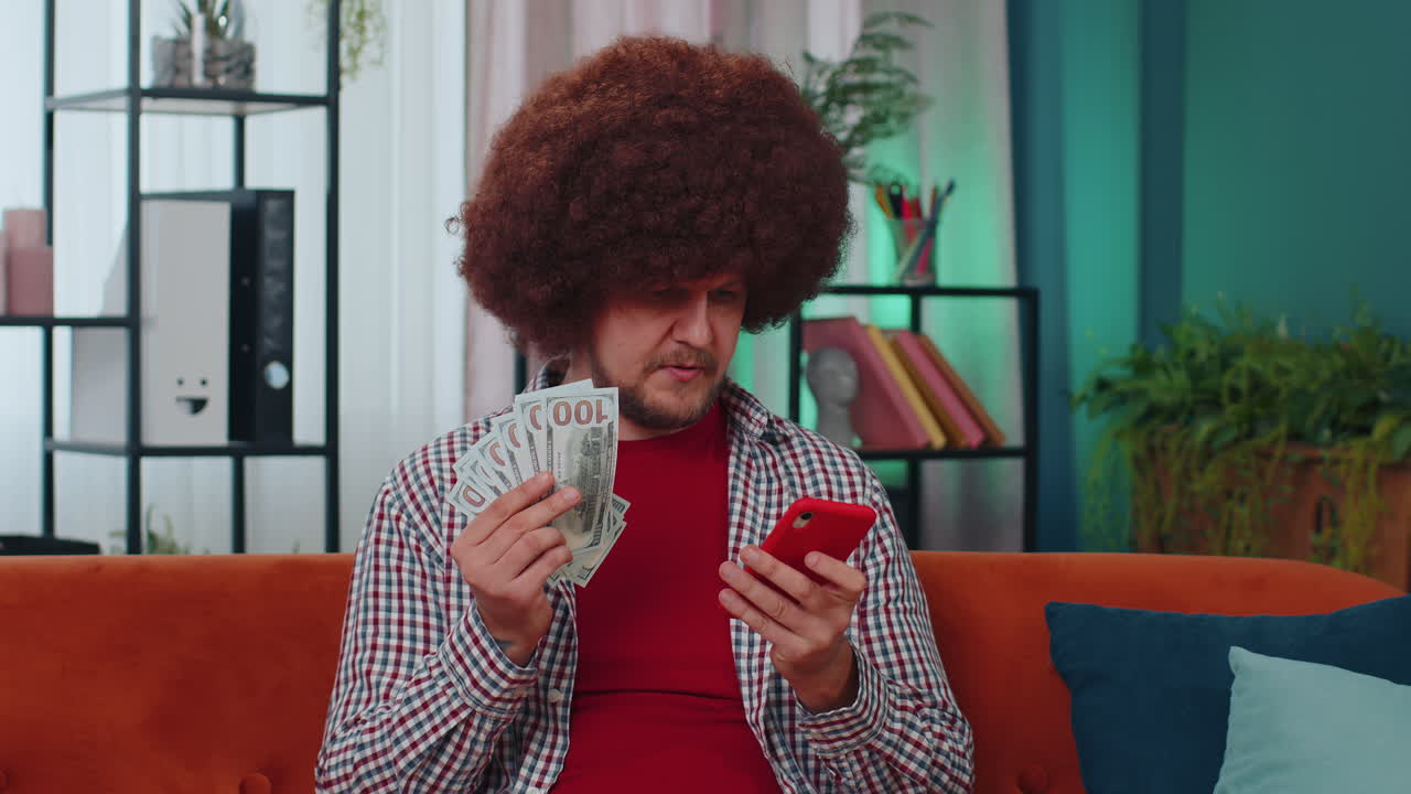 Smiling happy young adult man at home counting money cash use smartphone, income, saves, lottery win