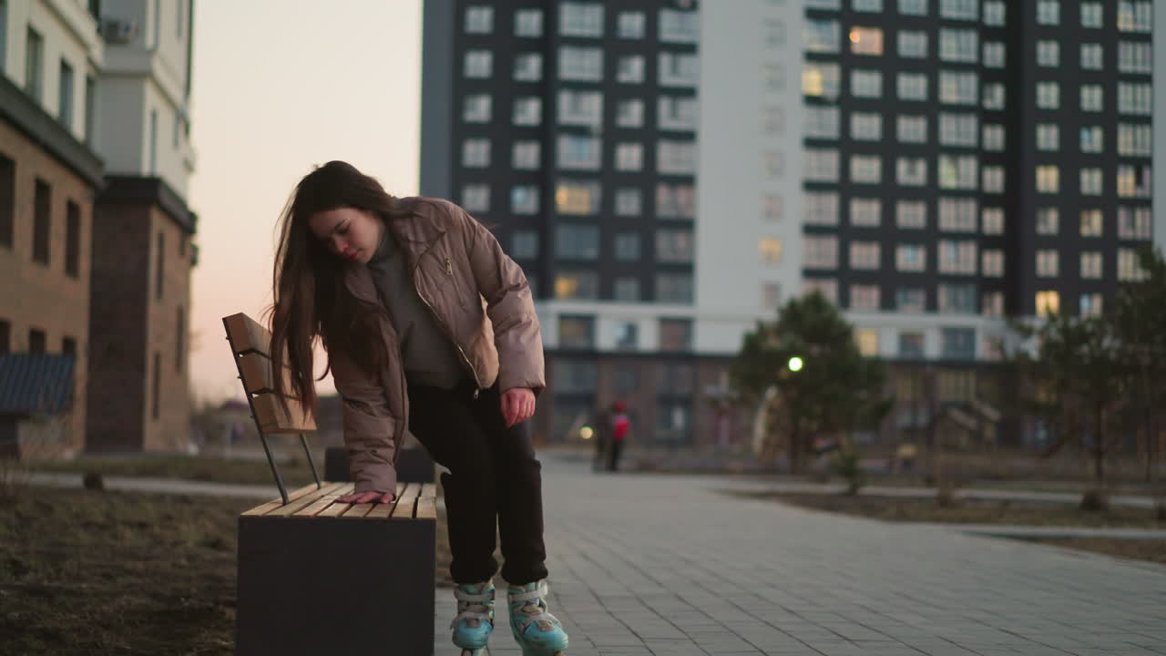 A girl in a peach jacket and black trousers skates towards a bench in an urban park. She bends forward, resting her head on her hand, appearing tired . The evening setting, buildings in the background