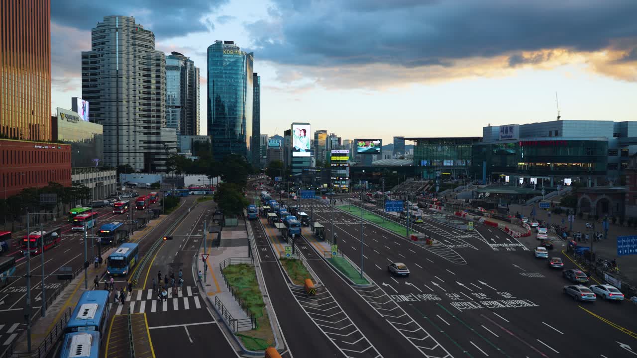 Seoul Station Bus Transfer Center at Sunset, People Crossing Road, Cars and Buses Traffic Moving With Towering Skyscrapers in Backdrop - Aerial pan right reveal