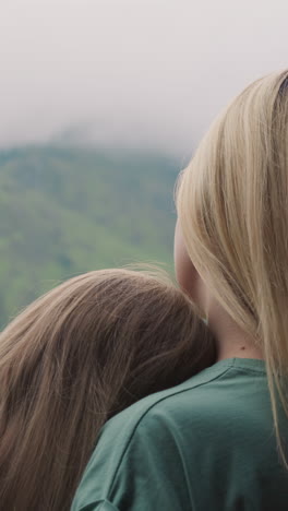 mother and adorable little daughter leaning on mom shoulder look at old distant mountains covered with deep mist in eco hotel close backside view slow motion