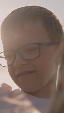 close-up of a young boy wearing glasses and a white shirt, standing beside his blurred mother. the boy points at the sky, his expression focused, capturing a curious and thoughtful moment outdoors