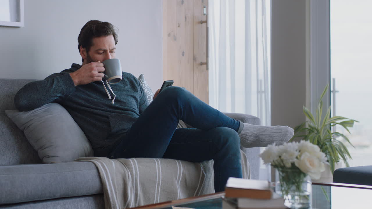 스마트폰을 사용하는 젊은 남자가 온라인 브라우징을 하고 소셜 미디어 메시지를 읽고 편안한 라이프 스타일을 즐기고 커피를 마시며 집에서 소파에서 휴식을 취하고 있습니다.