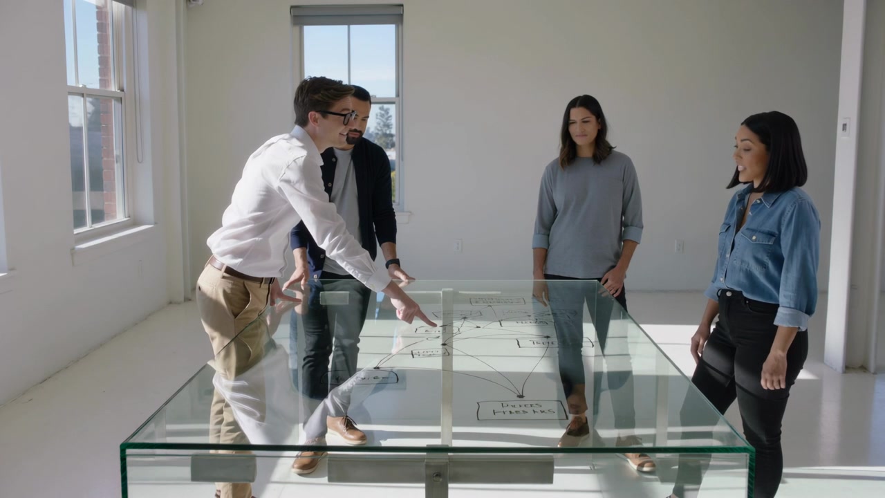 Hombre señalando, iniciando la revisión del estudio, inclinado y apuntando a un plano sobre cristal con un modelo, vestido con pantalones caqui.