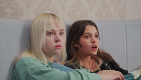 two young siblings sit side by side on bed watching movie with intense focus, one eating popcorn from bowl while both are wrapped in soft blankets