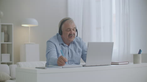hombre de mediana edad se está comunicando por internet usando computadora portátil y auriculares tomando y haciendo notas en el cuaderno