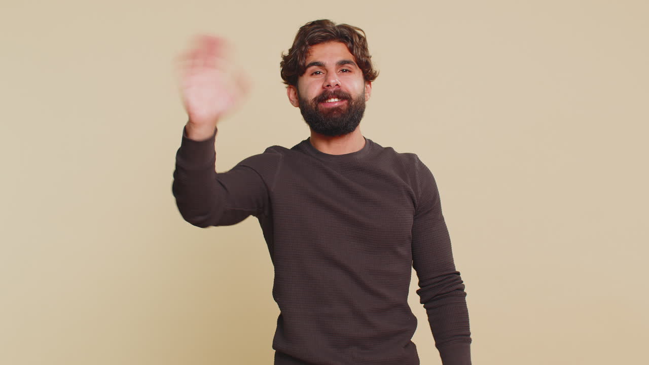 Handsome young indian man with long sleeves waving hello with his palm against beige background