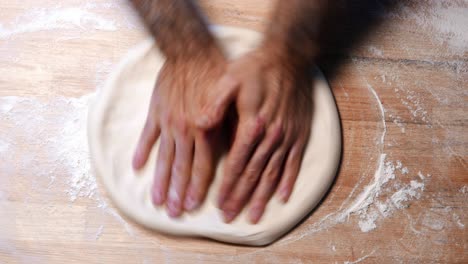 top-shot of hands of a skilled chef roll out the pizza dough on a wooden surface with flour around