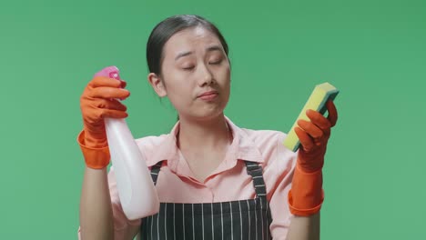 primer plano de una ama de casa asiática con un delantal tirando un spray y una esponja de cocina luego cruzando los brazos en el estudio de pantalla verde
