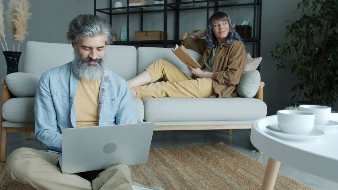 Couple relaxing at home, man working on laptop, woman reading a book