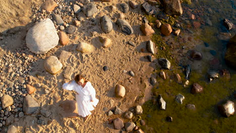 Aerial-Birds-Eye-View-Of-Women-Wearing-Flowing-White-Dress-Walking-Along-Rocky-Beach-Coastline-During-Golden-Hour