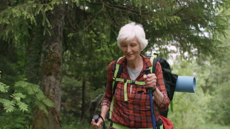 cheerful senior woman smiling while walking in forest with hiking backpack and trekking poles