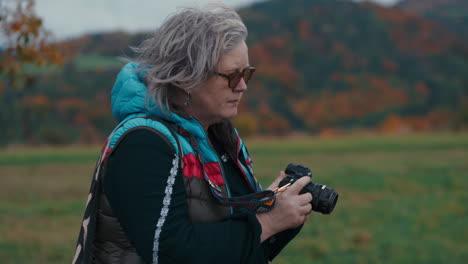 Close-up-shot-of-Female-Photographer-changing-aperture-in-her-camera,-pressing-a-shutter-and-taking-a-picture-of-a-beautiful-windy-autumn-nature-with-orange-leaves-in-the-background