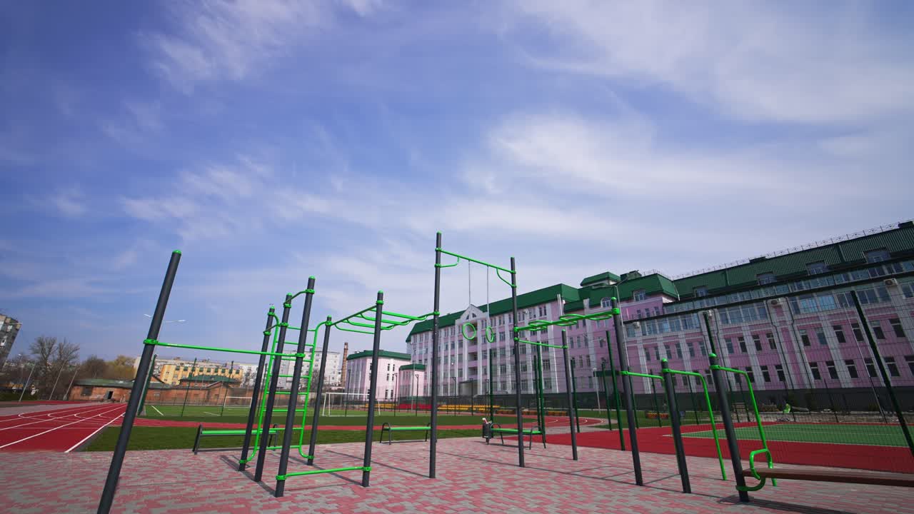 Approaching the set of sport horizontal bars at the outdoor sport ground with paved floor. Running tracks, football field and long pink building at backdrop.