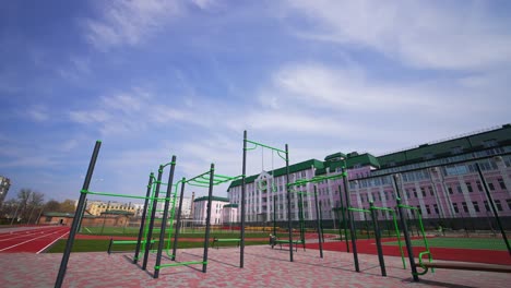 approaching the set of sport horizontal bars at the outdoor sport ground with paved floor. running tracks, football field and long pink building at backdrop.