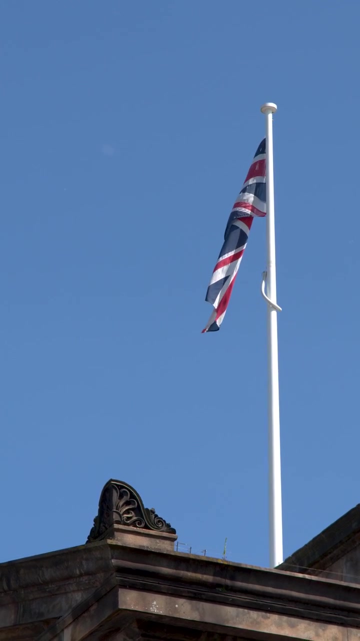 Bandeira Union Jack no topo de um edifício histórico de pedra sob um céu azul claro, luz solar do meio-dia, tomada estática.