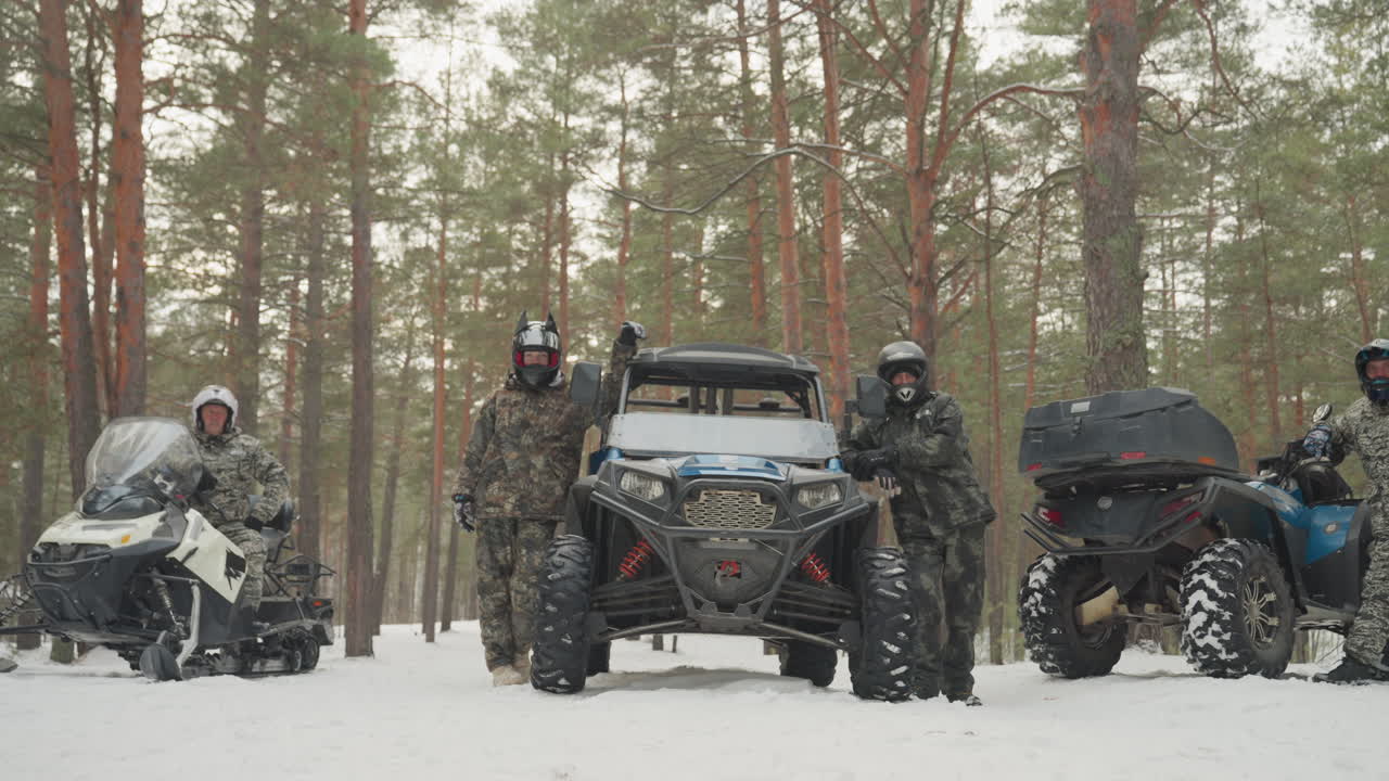 Schieramento nella foresta innevata con un convoglio di ATV e UTV. Un gruppo di piloti è in piedi accanto a un veicolo side-by-side e a una motoslitta in una silenziosa pineta, indossando equipaggiamento invernale mimetico, caschi e occhiali, con i motori al minimo.