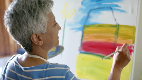 focused senior biracial woman wearing apron and painting on big canvas at home, slow motion