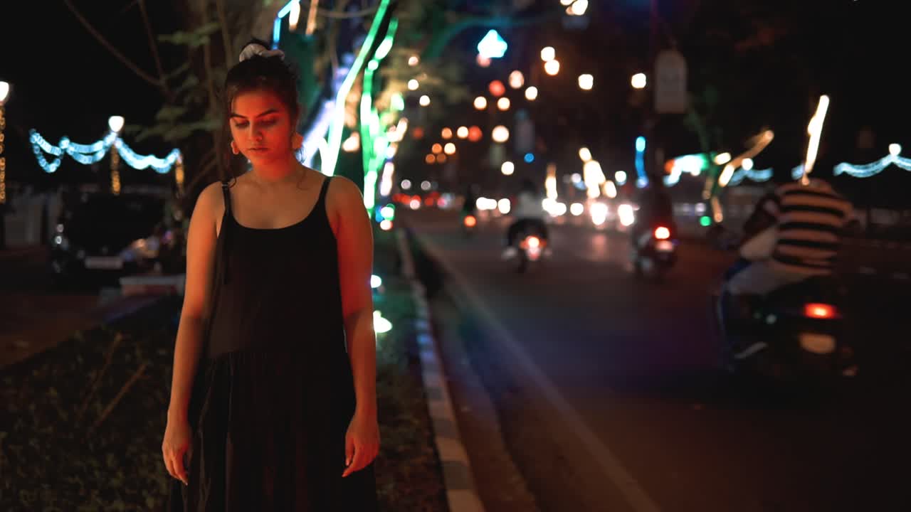 Ragazza indiana in abito nero in piedi tra le luci della città accanto a una strada con traffico intenso