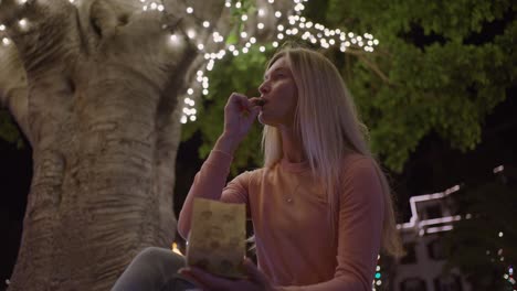 Attractive-woman-eating-freshly-roasted-chestnuts-from-paper-bag-under-string-light-filled-tree-at-night