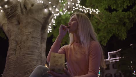 mujer atractiva comiendo castañas recién asadas de una bolsa de papel bajo un árbol lleno de luz por la noche