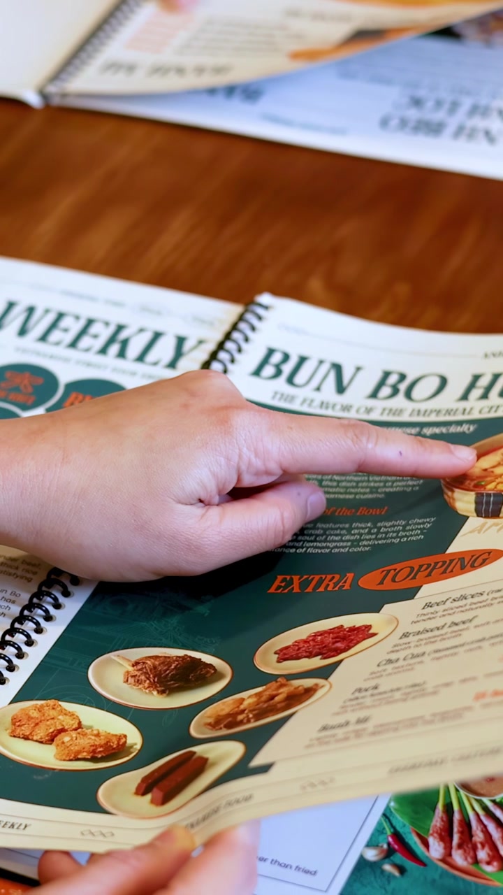 Nærbillede af en hånd, der peger på Bun Bo Hue på en restaurantmenu over træ.