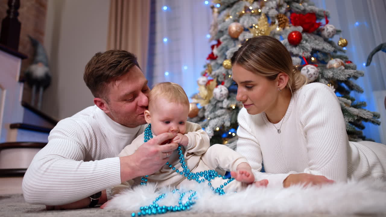 Loving mom and dad lie on the floor with their baby in the middle. Cute infant plays with beads decorations for the Christmas tree. Low angle view.