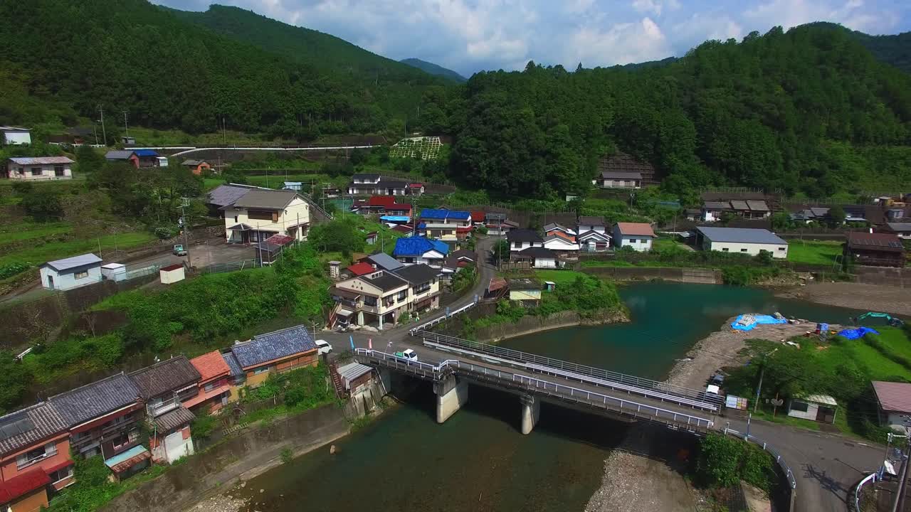 la vista serena del avión no tripulado viaja hacia adelante por encima de una pacífica aldea japonesa con un río cruzado por un puente situado en un valle aislado