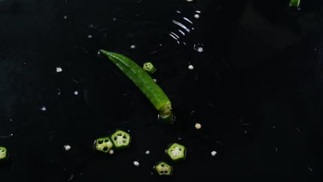 Okra-fruit-falling-on-wet-surface-in-black-studio