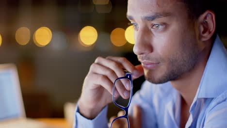 tired biracial businessman at desk with computers putting on glasses in office at night, slow motion