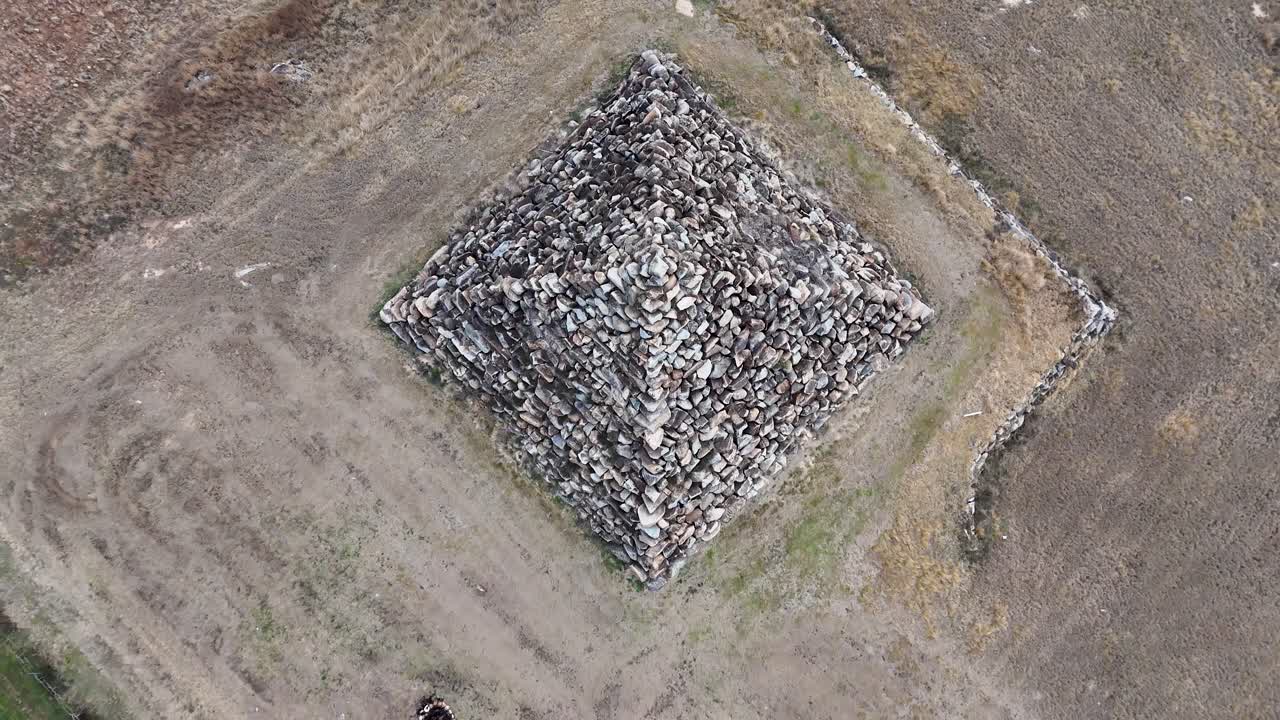 Drone footage captures a slow, smooth aerial rotation above a large stone pyramid structure in a dry, open landscape under natural daylight