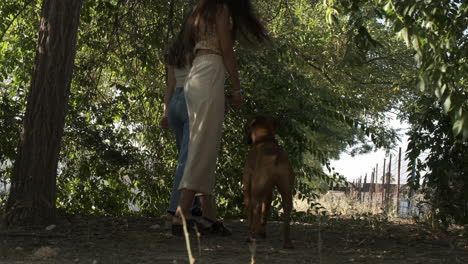 two young women and a dog walking outdoors in a natural setting