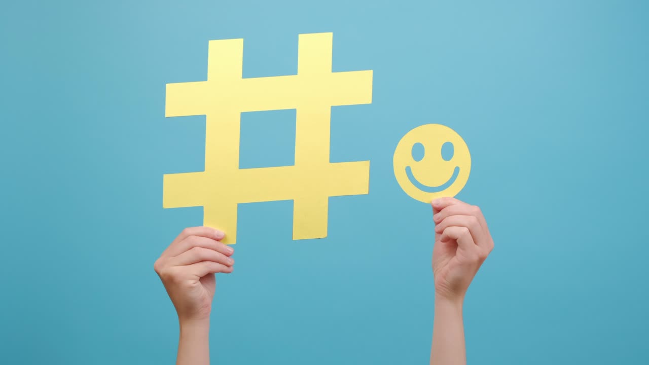 큰 노란색 해시태그 표지판과 작은 행복한 웃는 얼굴 아이콘을 들고있는 여성 손의 근접, 광고를 위해 복사 공간이있는 스튜디오의 파란색 배경 벽에 고립되어 있습니다.