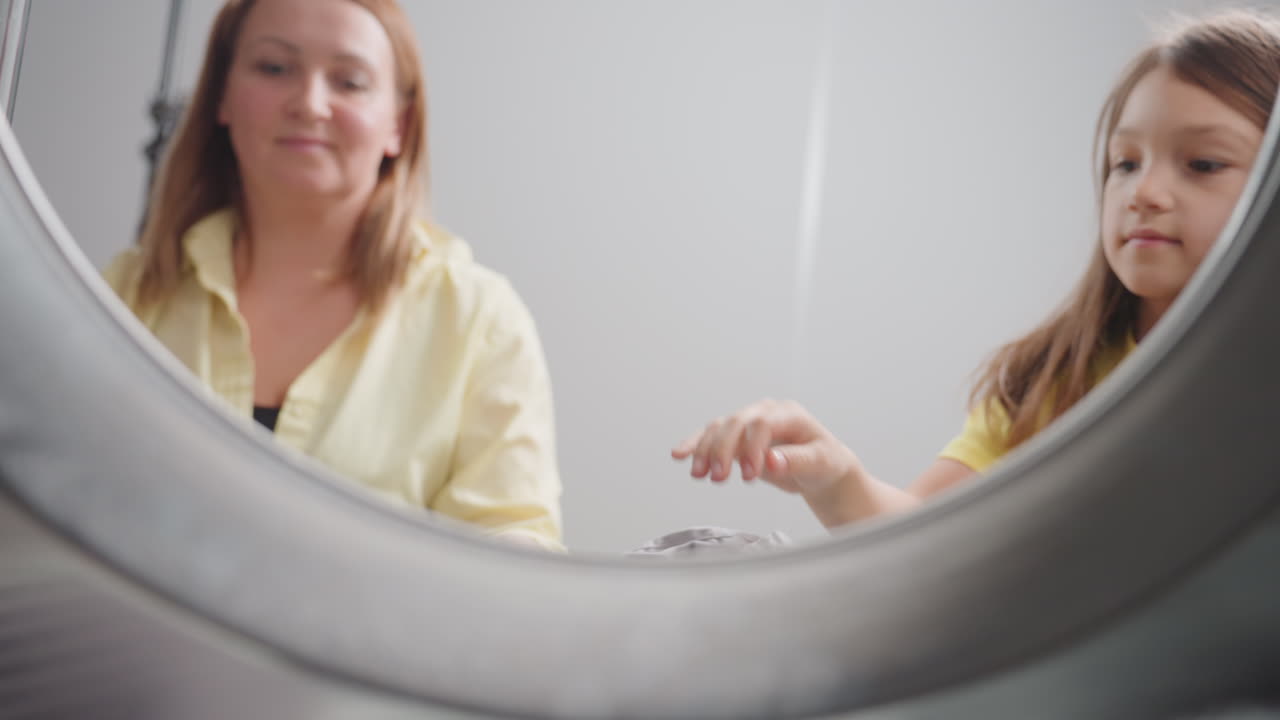 Inside washer pov shows business woman with energetic girl leaning in, looking at load before close, hands near drum rim, teamwork during laundry task, bright room, door closing moment