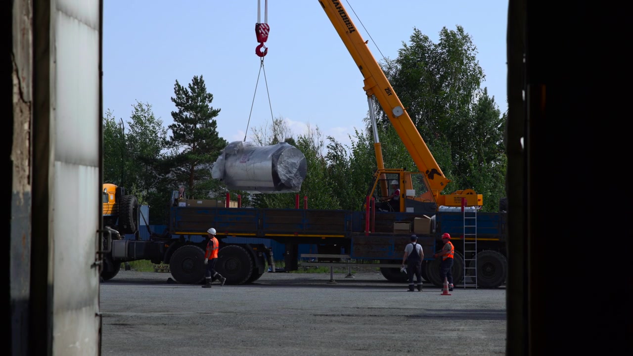Grúa móvil amarilla baja un gran tanque cilíndrico de metal envuelto en papel de aluminio reflectante sobre un camión de plataforma mientras el personal de tierra con equipo de seguridad guía la colocación precisa en un sitio industrial, con grúa, bajando