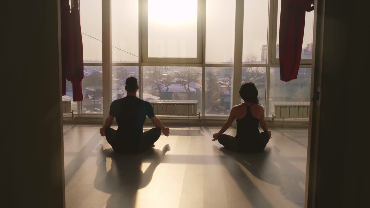 Un momento tranquillo di meditazione: due persone che praticano yoga in una stanza soleggiata con grandi finestre che si affacciano su un paesaggio suggestivo all'alba.