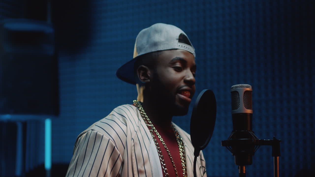 헤드폰을 끼고 어두운 사운드 스튜디오에서 노래를 녹음하는 모자 가수의 잘생기고 행복한 젊은 남자