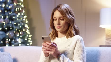 retrato cerrado de una mujer hermosa y alegre con cara sorprendida sentada en una habitación acogedora cerca de un árbol de navidad brillante escribiendo en un smartphone