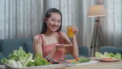 happy asian woman holding and pointing a glass of orange juice while having healthy food at home