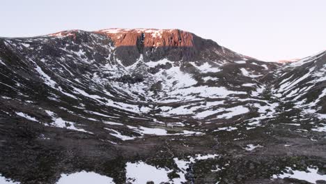 Aerial-drone-footage-slowly-reversing-to-reveal-sunrise-over-a-dramatic-vertical-cliff-rising-out-of-the-snow-and-moorland-in-winter-with-Hutchison-Memorial-Bothy,-Glen-Derry,-Cairngorms-National-Park