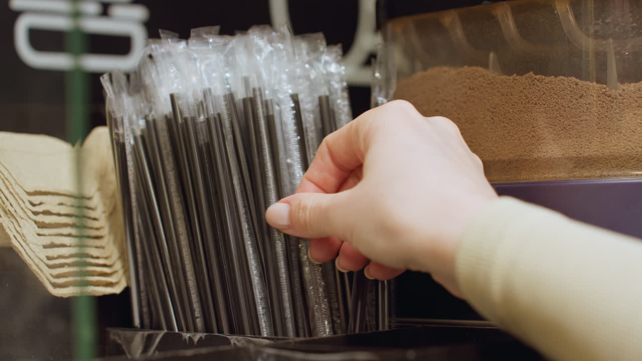 shopper hand selecting straw at counter, paper wrapped straws in clear dispenser, closeup of careful selection, warm cafe lighting, morning beverage station, focus on hygiene and packaging options