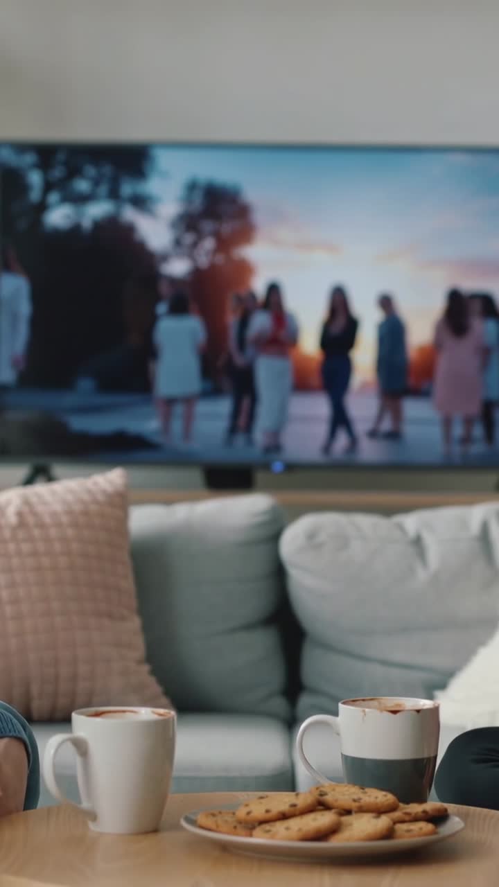 Video verticale: Inquadratura di un tavolino da caffè, due tazze e biscotti a casa, TV che mostra donne che ballano, jeans.