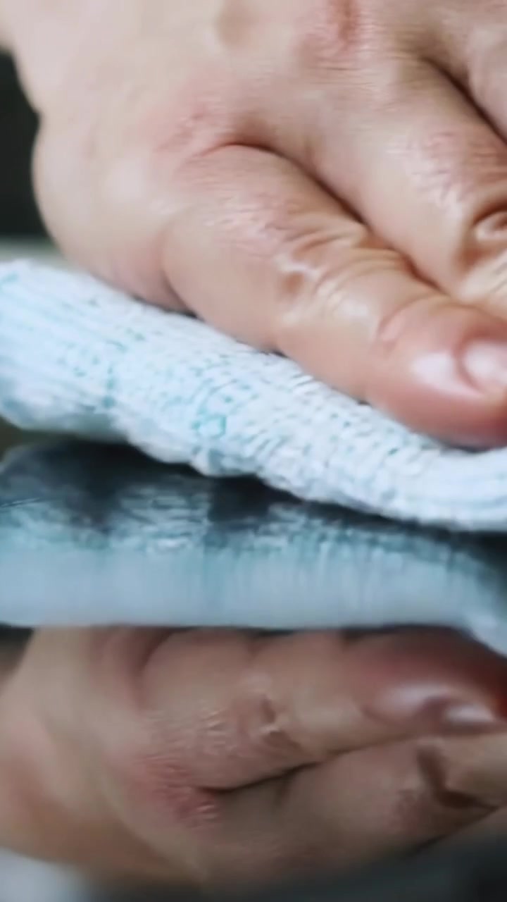 Video verticale: Posizionando una mano con un polsino blu, premendo e pulendo un panno blu piegato per lucidare il tavolo.