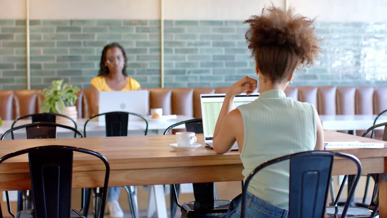 Donne afroamericane che lavorano sui laptop a un tavolo comune, con la donna in primo piano che si sfrega il collo mentre digita.